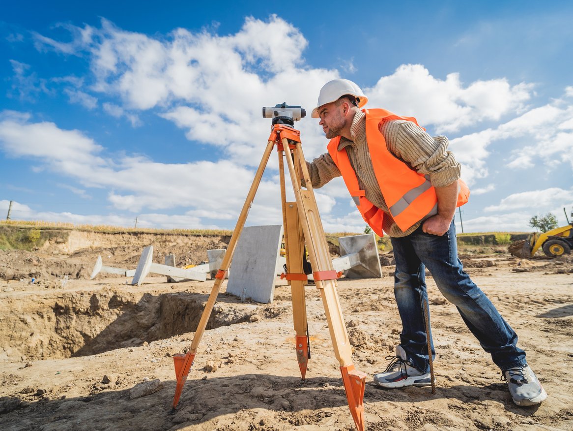 Vermessungsingenieur in reflektierender Sicherheitsweste bedient ein Theodolit-Vermessungsinstrument auf einer Baustelle mit sichtbaren Erdarbeiten und Baumaschinen im Hintergrund.