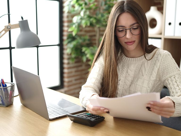 Junge Frau mit Brille sitzt an einem Schreibtisch, liest ein Dokument und arbeitet an einem Laptop, mit einer Schreibtischlampe und einem Taschenrechner neben sich.