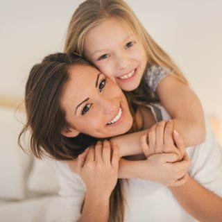 Eine strahlende Mutter und ihre fröhliche Tochter umarmen sich liebevoll, beide lächelnd und in die Kamera blickend. Ihre Freude und enge Bindung sind in dieser herzlichen Szene zu Hause deutlich spürbar.