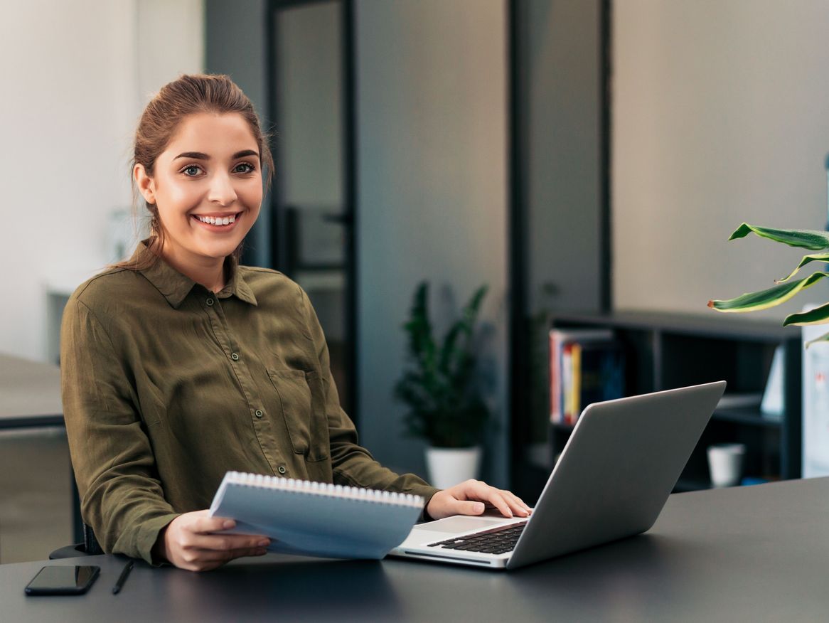 Eine Frau mit einem freundlichen Lächeln sitzt an ihrem Arbeitsplatz vor einem Laptop und hält Dokumente in der Hand. Ihre professionelle Haltung und das moderne Büro im Hintergrund spiegeln eine angenehme Arbeitsumgebung wider.