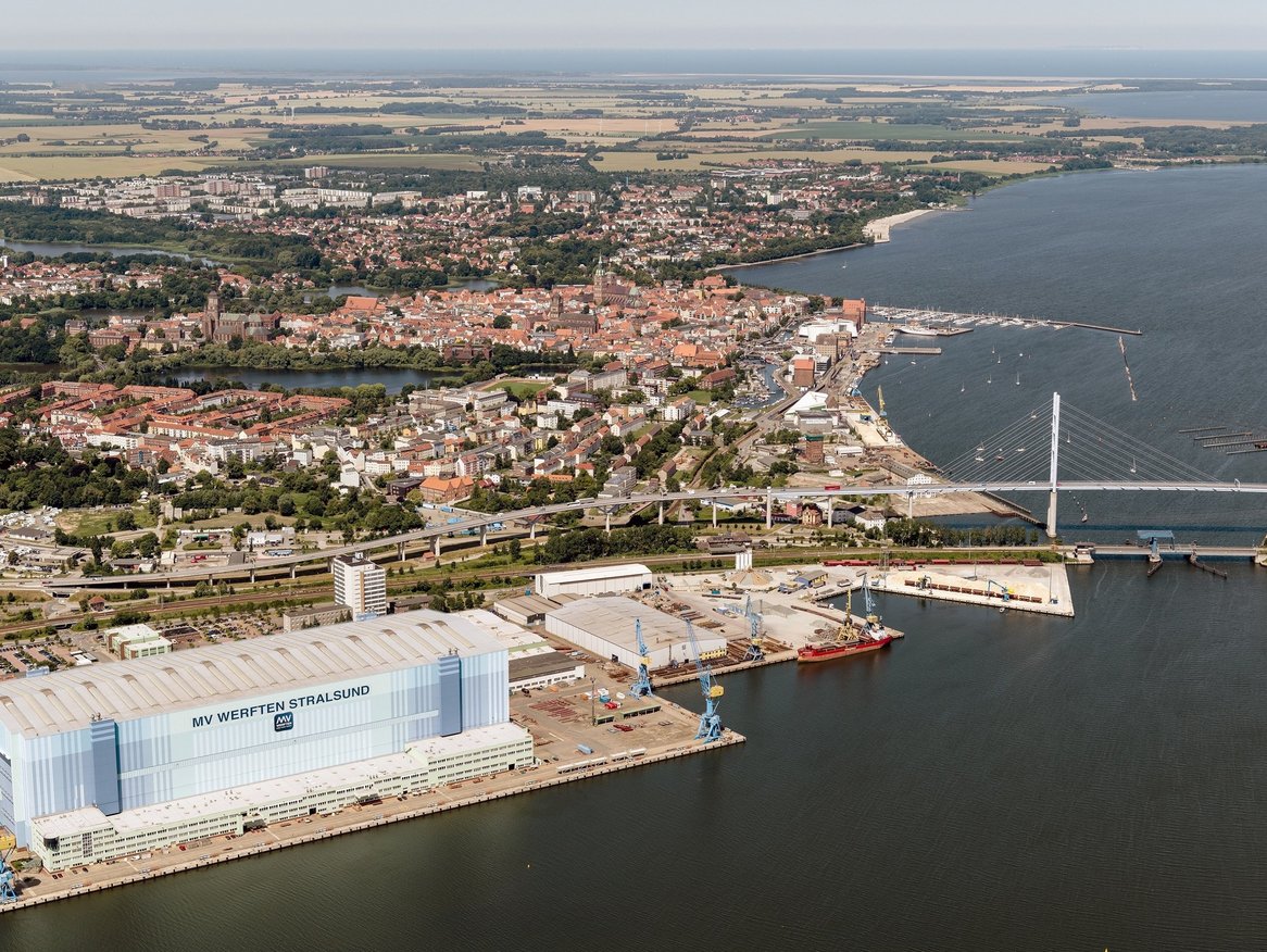Eine Luftaufnahme von Stralsund mit dichter Bebauung, die bis an das Ufer eines großen Gewässers reicht, mit einer Brücke im Hintergrund, die zwei Uferseiten verbindet.