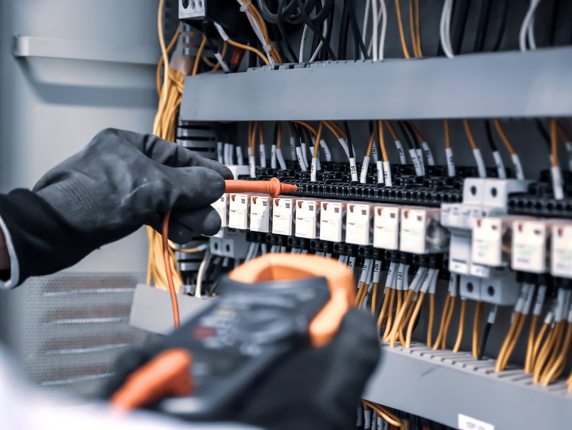 Detailaufnahme der Hände eines Elektrikers in Schutzhandschuhen, der an einer Schalttafel mit Kabeln und Sicherungselementen arbeitet.