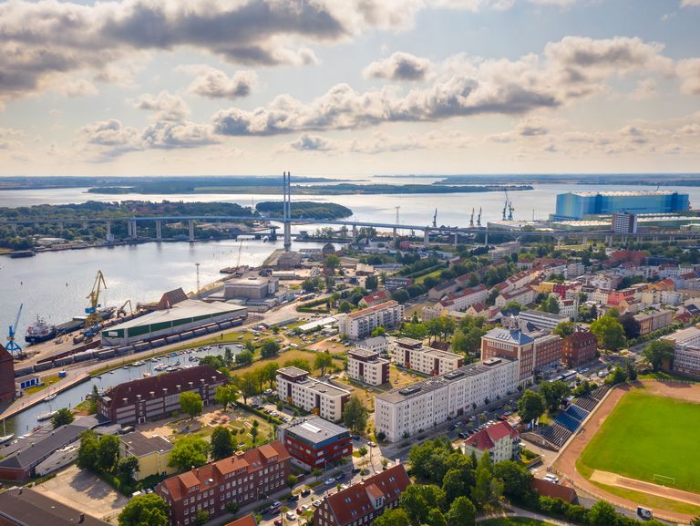 Ein Luftbild zeigt eine Stadt am Wasser, mit einer Brücke, die zwei Ufer verbindet, und industriellen Strukturen im Hintergrund. Die Mischung aus Wohnvierteln, Gewässern und industriellen Anlagen gibt einen Eindruck von der Vielfalt städtischer Umgebungen.