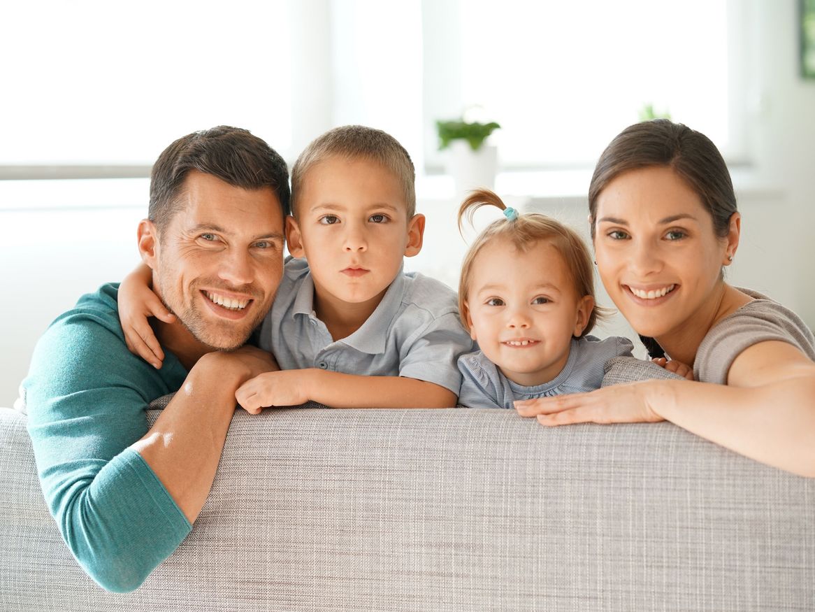 Eine glückliche Familie teilt einen Moment des Lächelns vor der Kamera, wobei die Eltern liebevoll ihre beiden jungen Kinder umarmen. Der Vater in einem türkisfarbenen Sweatshirt und die Mutter in einem grauen Top flankieren den ernst blickenden Sohn und die lächelnde Tochter mit einer hellblauen Schleife im Haar.