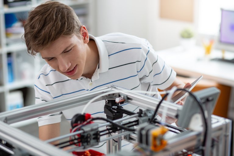 Junger Mann in einem gestreiften Hemd, der konzentriert an einem 3D-Drucker arbeitet, in einem hellen Raum mit Computer im Hintergrund.