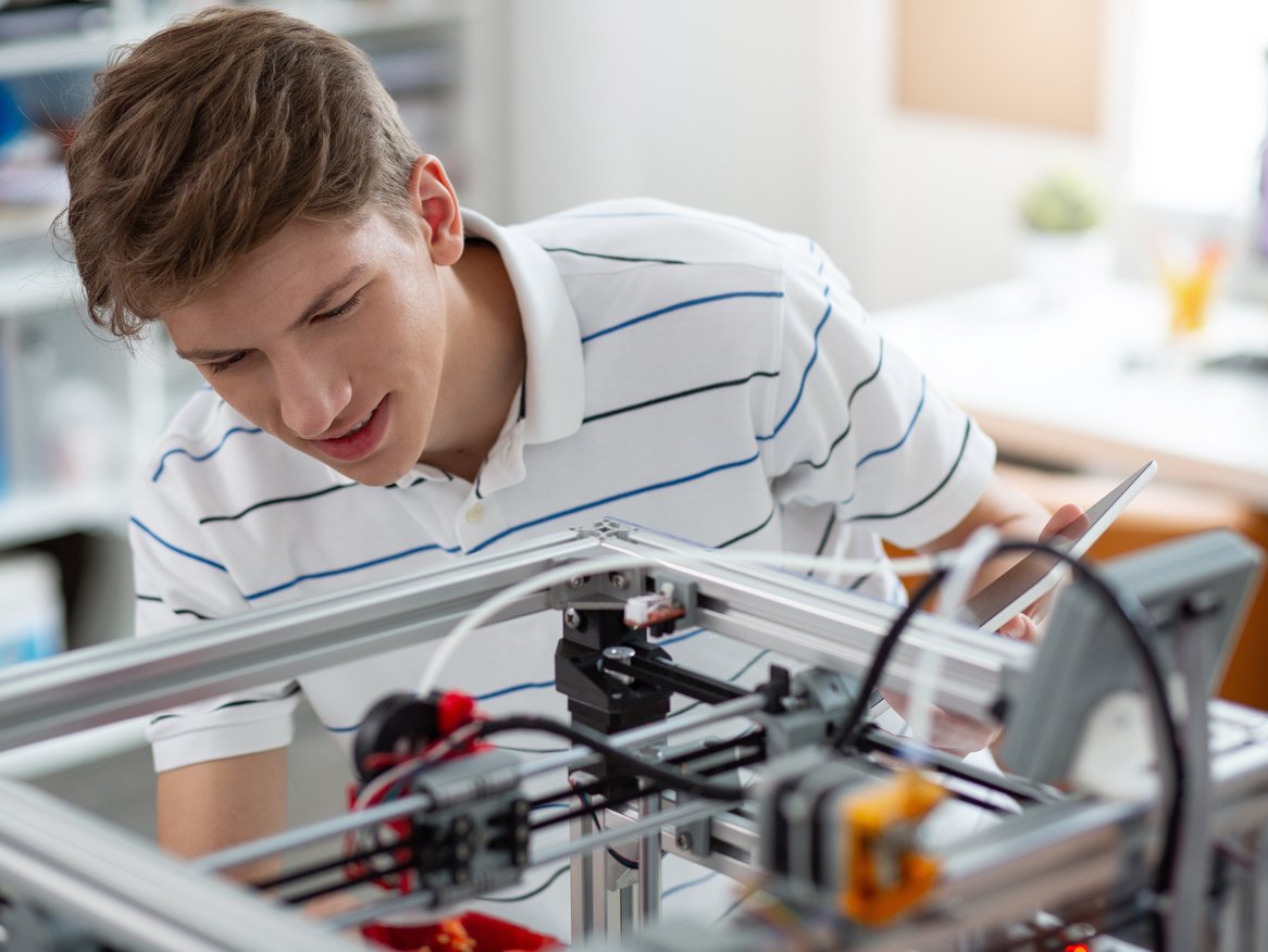 Junger Mann in einem gestreiften Hemd, der konzentriert an einem 3D-Drucker arbeitet, in einem hellen Raum mit Computer im Hintergrund.