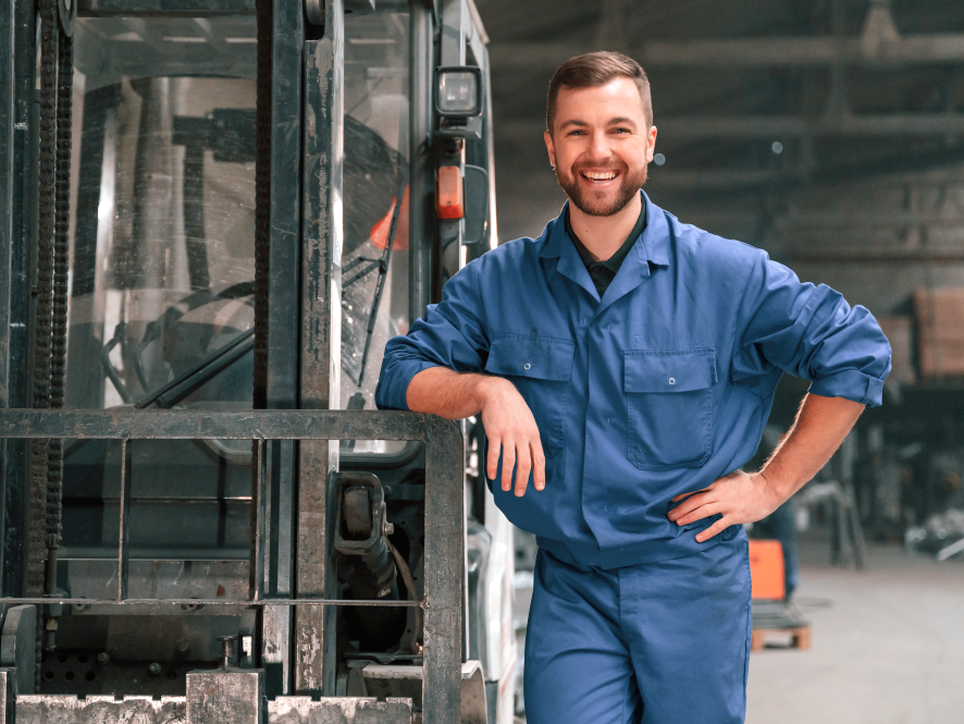 Ein lächelnder Mitarbeiter in blauer Arbeitskleidung steht neben einem Gabelstapler in einer industriellen Lagerhalle.
