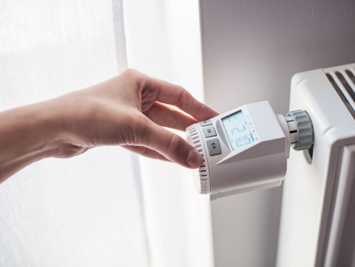 Eine Hand stellt die Temperatur an einem digitalen Thermostat eines Heizkörpers ein, was die alltägliche Handhabung von Heizsystemen in einem Haushalt und das Bemühen um Energieeffizienz widerspiegelt.