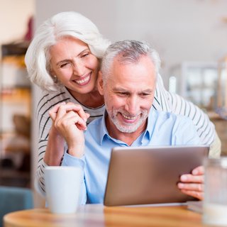 Ein älteres Paar, sichtlich in trauter Zweisamkeit, schaut gemeinsam auf ein Tablet. Sie lächeln fröhlich und verbunden, ein Bild häuslicher Harmonie und moderner Techniknutzung im Alter.