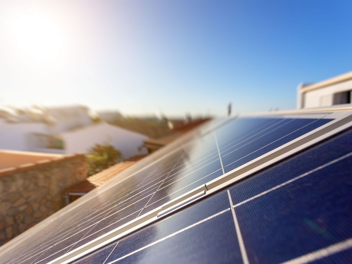 Das Foto zeigt die Sonne, die auf ein Solarpanel auf dem Dach eines Hauses mit blauem Himmel scheint.