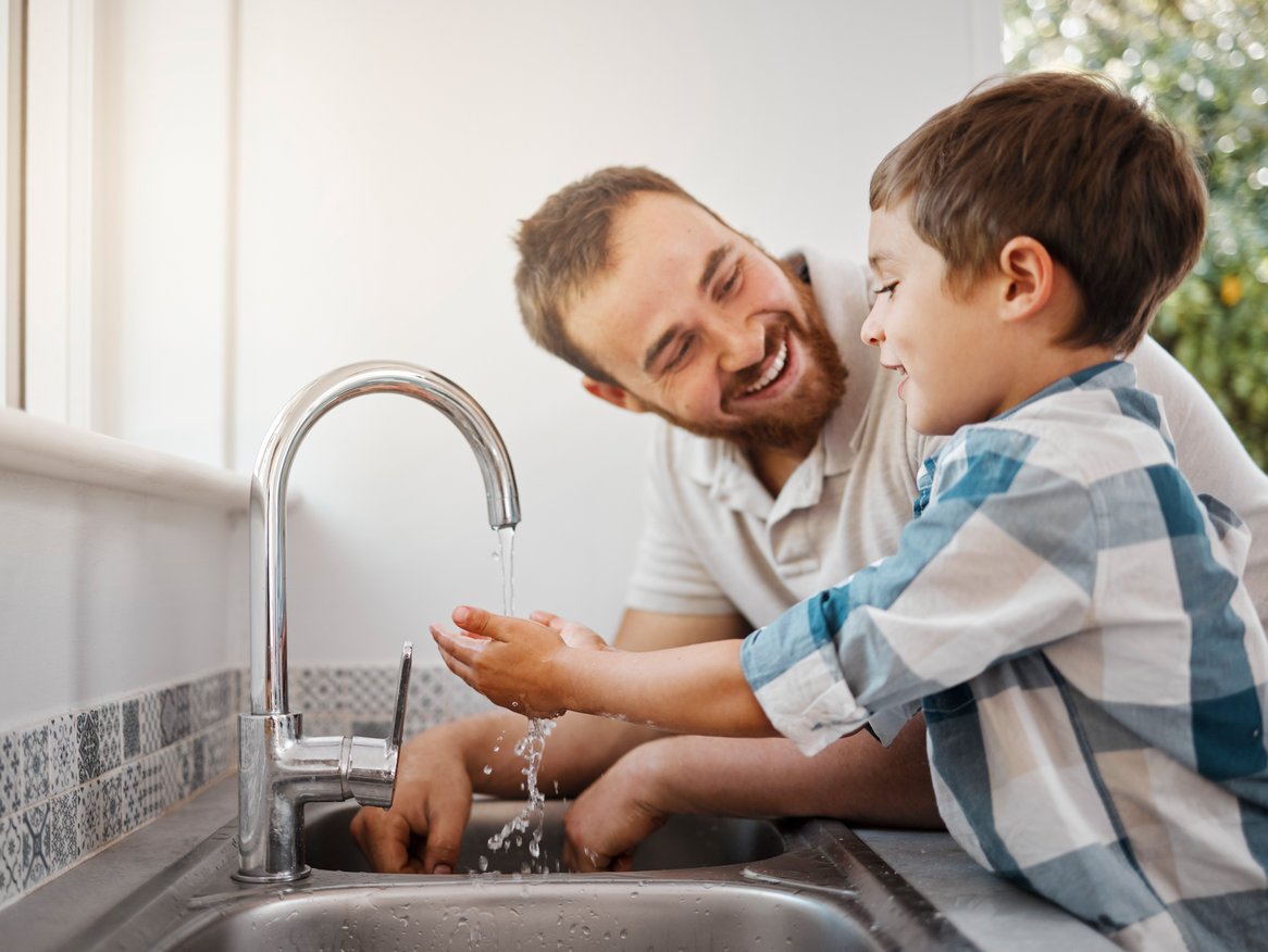Ein herzlicher Moment zwischen Vater und Sohn beim Händewaschen in der Küche. Beide lachen sich an, während sie spielerisch das Wasser aus dem Hahn spritzen, was die Freude an gemeinsamen alltäglichen Aktivitäten und die Eltern-Kind-Bindung betont.