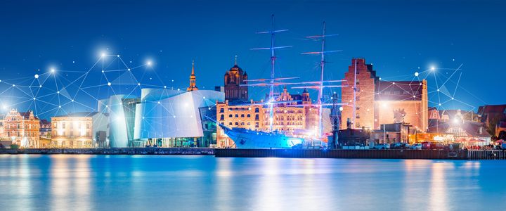 Eine Panoramaansicht einer beleuchteten Hafenstadt bei Nacht, mit einem Schiff im Vordergrund und einer Verbindung aus Lichtpunkten über dem Stadtbild, was auf Vernetzung und digitale Technologie hinweist.