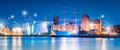 Eine Panoramaansicht einer beleuchteten Hafenstadt bei Nacht, mit einem Schiff im Vordergrund und einer Verbindung aus Lichtpunkten über dem Stadtbild, was auf Vernetzung und digitale Technologie hinweist.