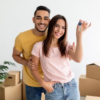 Ein glückliches Paar posiert mit einem Schlüssel in einer neuen Wohnung, umgeben von Umzugskartons, lächelnd und freudig über den neuen Lebensabschnitt.