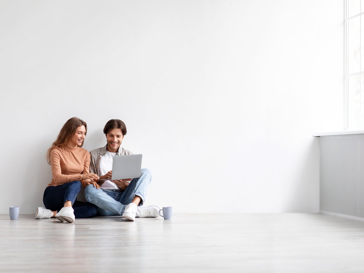 Ein junges Paar sitzt entspannt auf dem Boden eines minimalistisch eingerichteten Raumes, vertieft in ein Gespräch mit einem Laptop zwischen ihnen. Das Bild strahlt Ruhe und moderne Einfachheit aus und könnte das Konzept von Work-Life-Balance oder gemeinsamer Planung zu Hause darstellen.