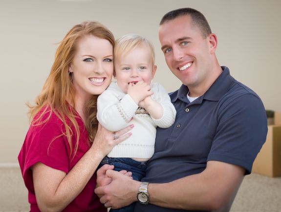 Eine glückliche Familie, bestehend aus Mutter, Vater und Baby, sitzt in einem fast leeren Raum mit Umzugskartons im Hintergrund, was auf einen neuen Lebensabschnitt im eigenen Zuhause hinweist. Die Wärme und Freude des Zusammenseins kommen deutlich zum Ausdruck.