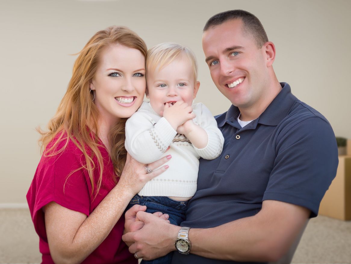 Eine glückliche Familie, bestehend aus Mutter, Vater und Baby, sitzt in einem fast leeren Raum mit Umzugskartons im Hintergrund, was auf einen neuen Lebensabschnitt im eigenen Zuhause hinweist. Die Wärme und Freude des Zusammenseins kommen deutlich zum Ausdruck.