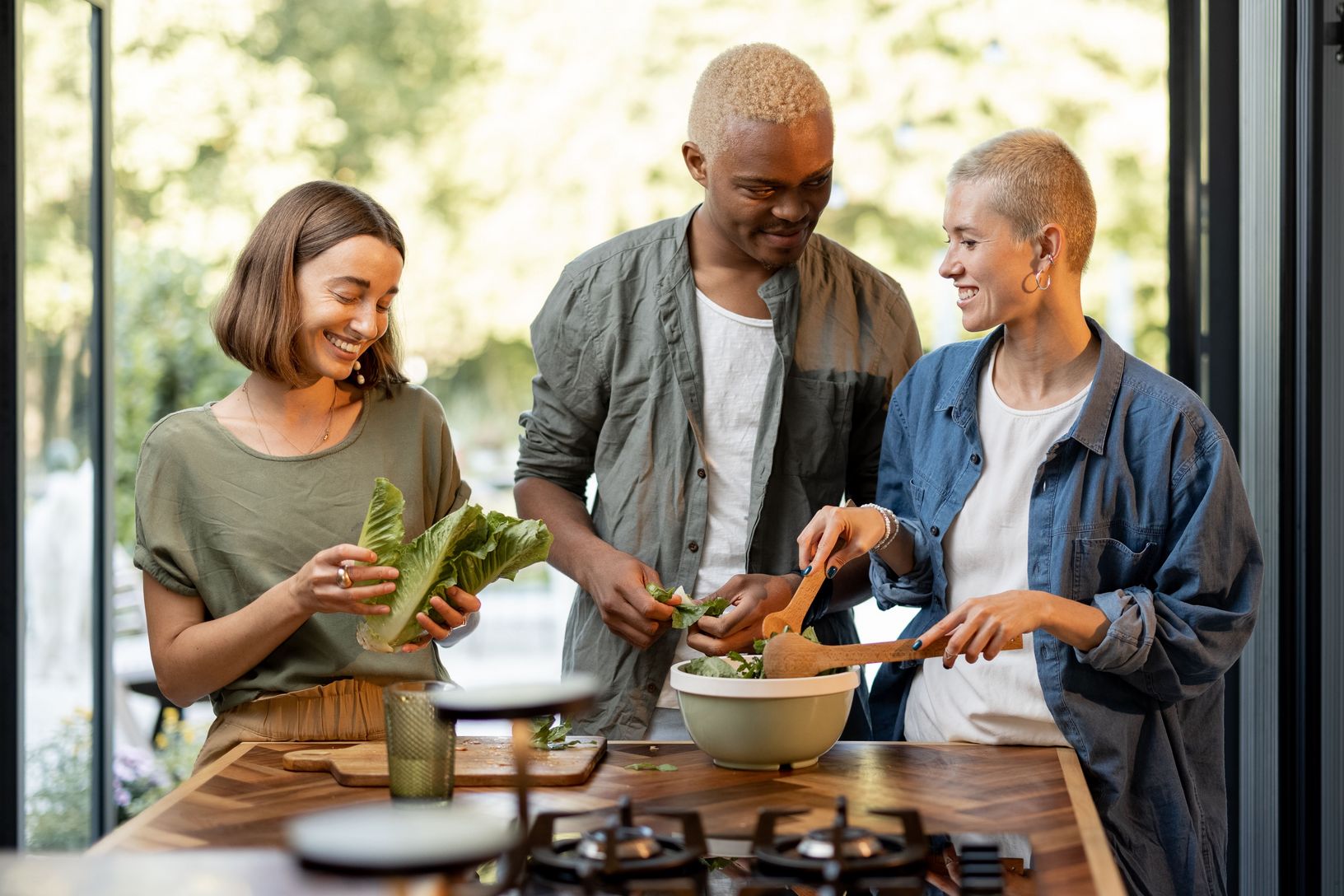 Eine Familie teilt einen gemeinsamen Moment beim Kochen in der Küche. Ein älterer Mann und eine Frau, wahrscheinlich ein Paar, stehen zusammen mit einer jüngeren Frau am Herd, während sie gemeinsam an der Zubereitung einer Mahlzeit arbeiten. Sie scheinen entspannt und in ihre Aufgabe vertieft, was auf eine warme und familiäre Atmosphäre hinweist.