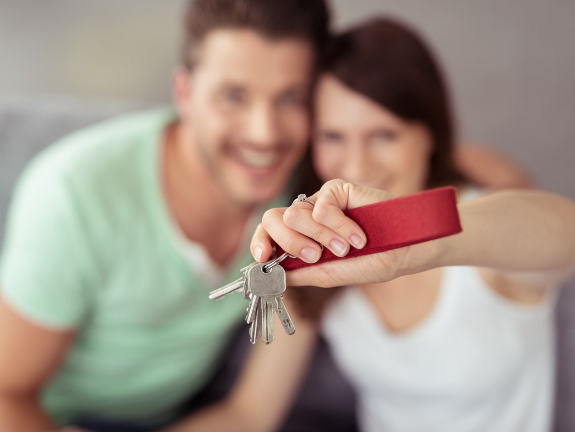 Ein unscharfes Paar im Hintergrund hält gemeinsam einen Schlüsselbund mit einem roten Schlüsselanhänger in den Fokus der Kamera, ein Symbol für neue Hausbesitzer, die den Schlüssel zu ihrem neuen Zuhause präsentieren.