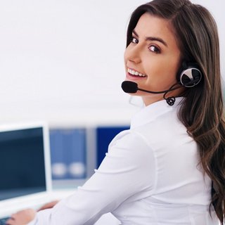 Eine freundlich lächelnde Frau im Kundendienst mit Headset blickt zurück, während sie an ihrem Arbeitsplatz vor einem Computer sitzt. Ihre professionelle Haltung und die moderne Büroumgebung deuten auf einen kundenorientierten Service hin.