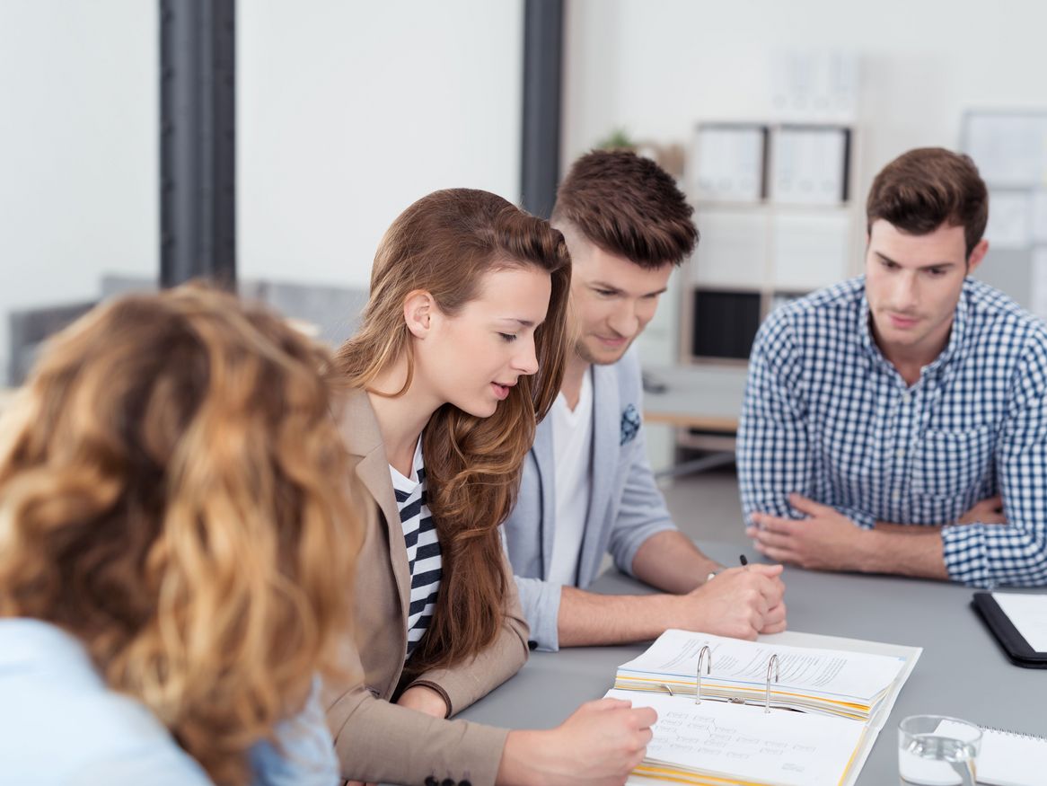 Vier junge Erwachsene in einem Büro, die sich auf Unterlagen konzentrieren, die auf einem Tisch ausgebreitet sind, mit einem unscharfen Vordergrund.