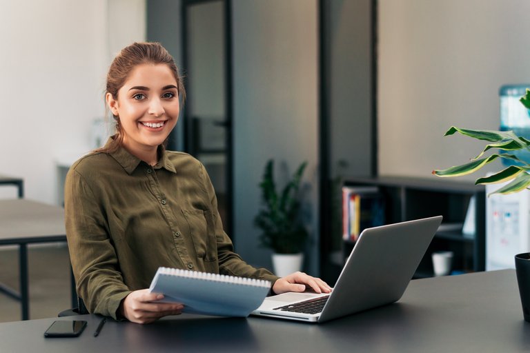 Eine Frau mit einem freundlichen Lächeln sitzt an ihrem Arbeitsplatz vor einem Laptop und hält Dokumente in der Hand. Ihre professionelle Haltung und das moderne Büro im Hintergrund spiegeln eine angenehme Arbeitsumgebung wider.