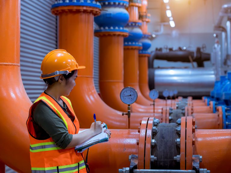 Industriearbeiterin in Schutzausrüstung notiert Messwerte an einer Rohrleitung in einer Anlage mit großen orangen und blauen Rohren.