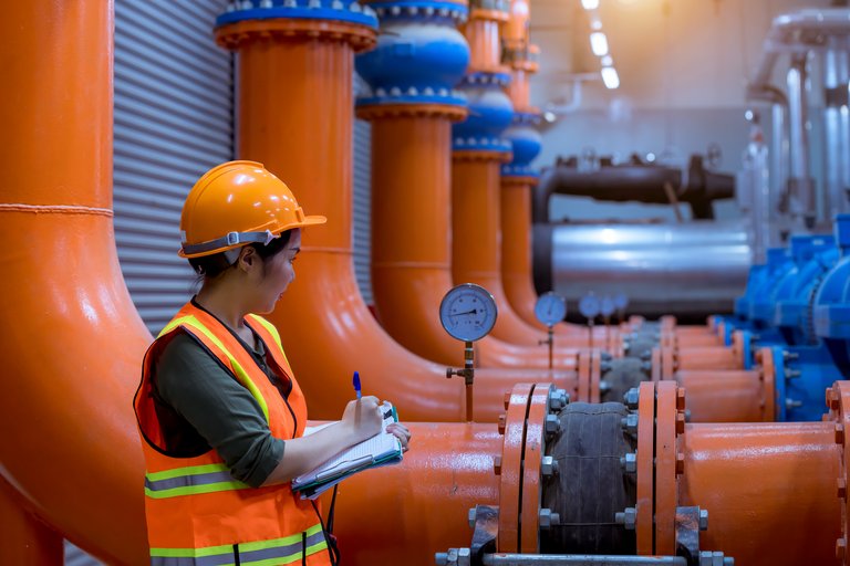 Industriearbeiterin in Schutzausrüstung notiert Messwerte an einer Rohrleitung in einer Anlage mit großen orangen und blauen Rohren.