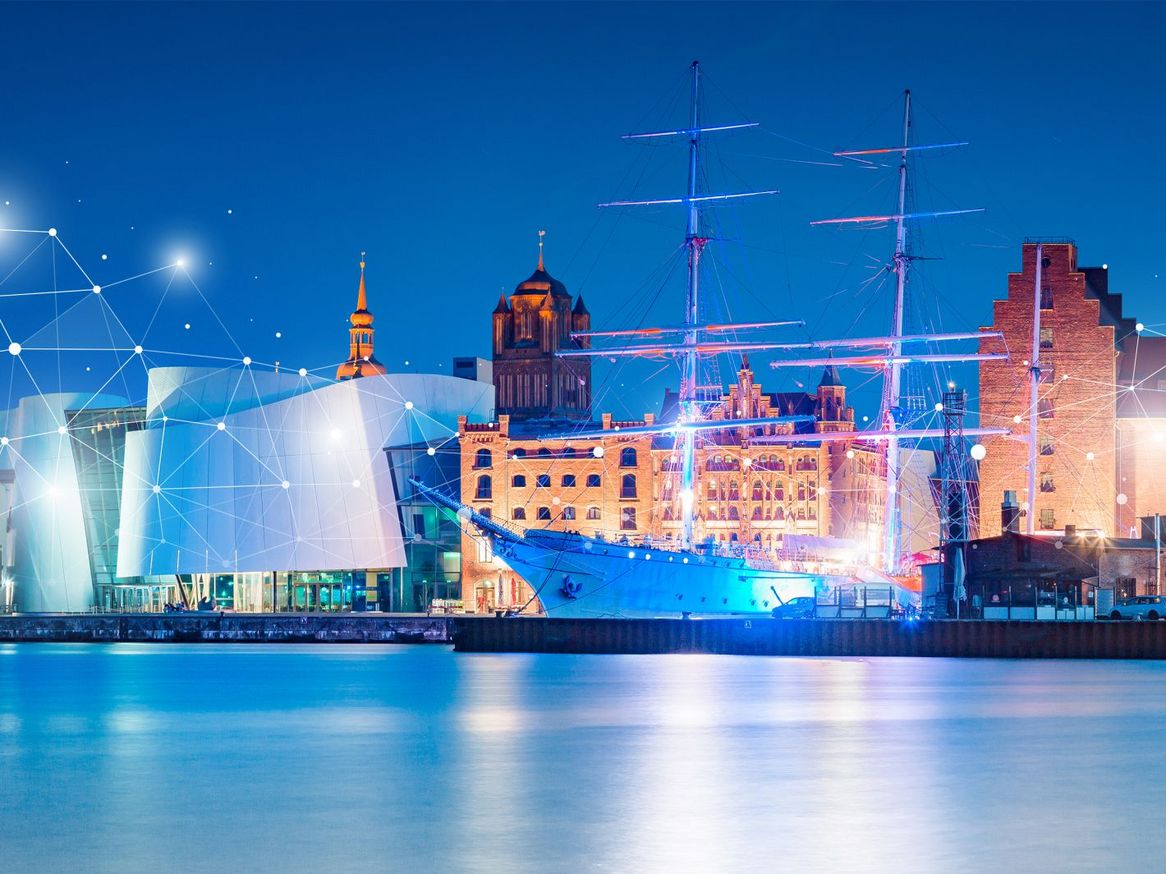 Eine Panoramaansicht einer beleuchteten Hafenstadt bei Nacht, mit einem Schiff im Vordergrund und einer Verbindung aus Lichtpunkten über dem Stadtbild, was auf Vernetzung und digitale Technologie hinweist.