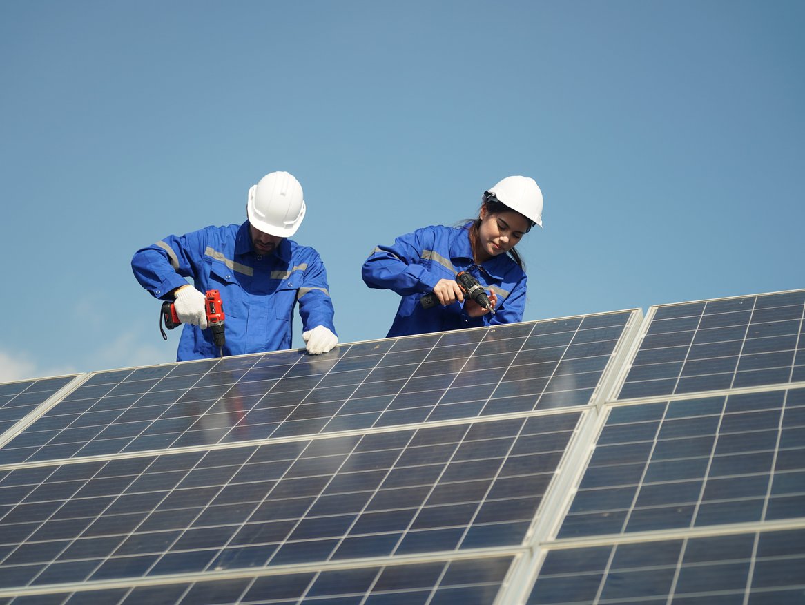 Zwei Techniker in blauer Arbeitskleidung und Schutzhelmen installieren Solarpaneele auf einem Dach, unterstreichen die Arbeit an nachhaltiger Energie.