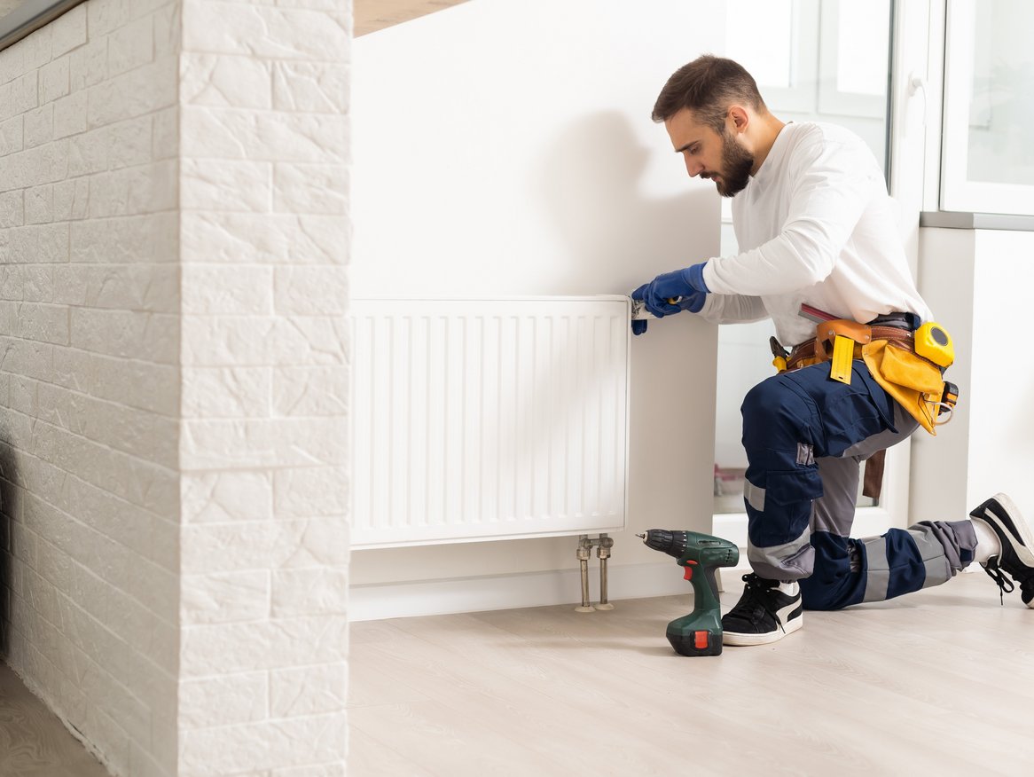 Heizungsmonteur in Arbeitskleidung repariert einen weißen Heizkörper in einem modernen Raum mit weißer Wand und Holzboden.