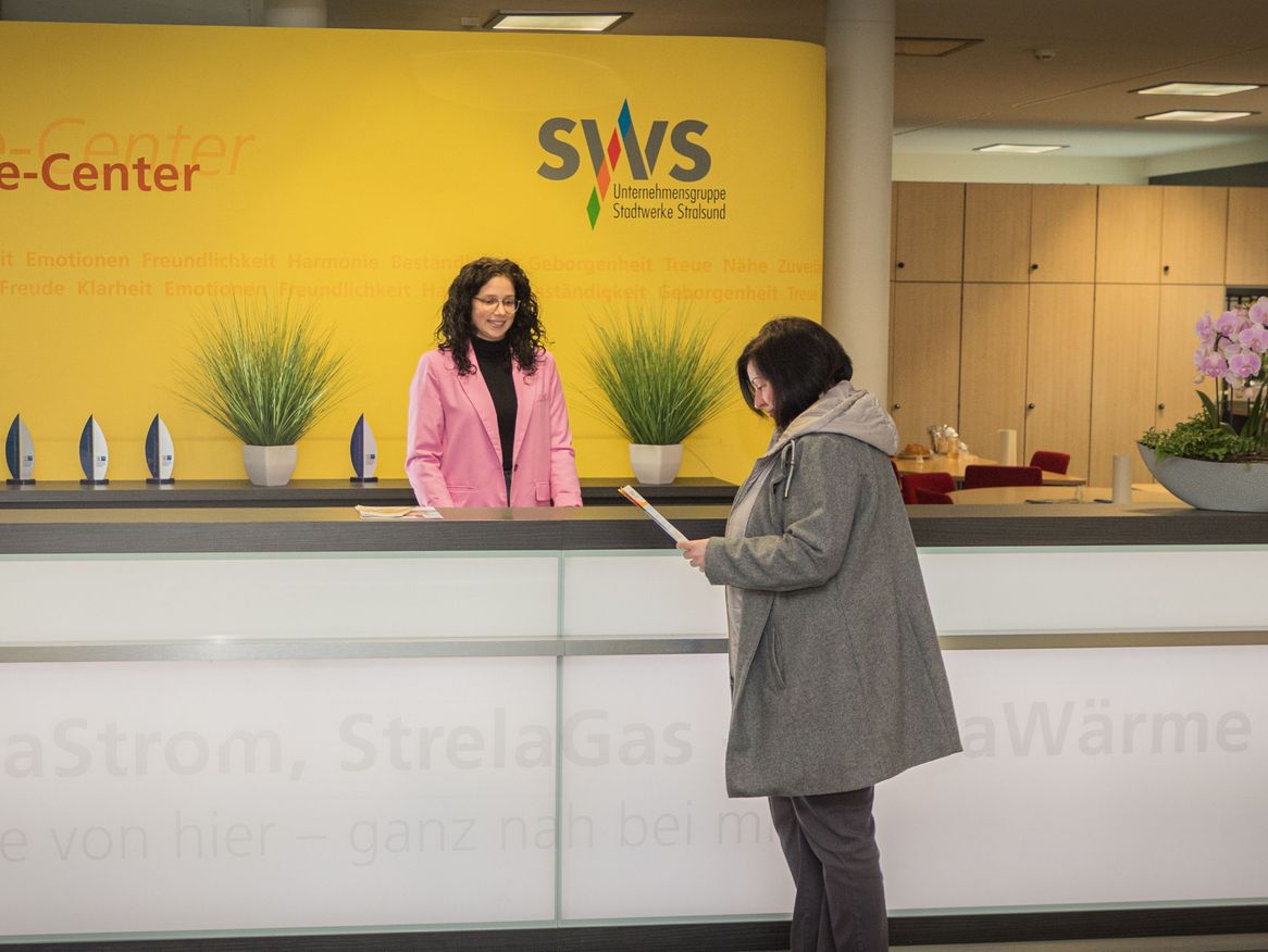 Ein angenehmer Service-Moment wird im Service-Center der Stadtwerke Stralsund eingefangen, wo eine Kundin in einer grauen Jacke am Empfang von einer Mitarbeiterin mit lockigem Haar und einem leuchtend rosa Mantel bedient wird. Im Hintergrund ist eine gelbe Wand mit den Werten des Unternehmens, wie Beständigkeit und Zuverlässigkeit, zu sehen