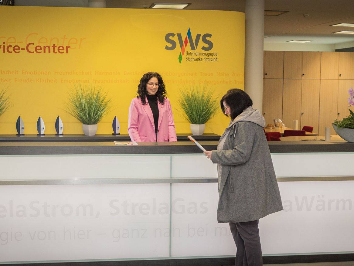 Ein angenehmer Service-Moment wird im Service-Center der Stadtwerke Stralsund eingefangen, wo eine Kundin in einer grauen Jacke am Empfang von einer Mitarbeiterin mit lockigem Haar und einem leuchtend rosa Mantel bedient wird. Im Hintergrund ist eine gelbe Wand mit den Werten des Unternehmens, wie Beständigkeit und Zuverlässigkeit, zu sehen