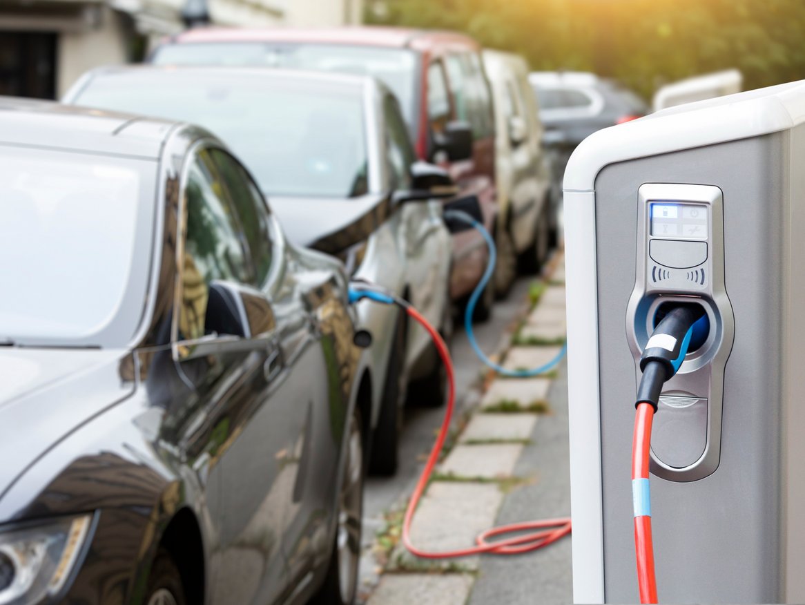 Eine moderne Ladestation für Elektrofahrzeuge mit angeschlossenem Ladekabel, das ein geparktes Elektroauto lädt, verschwommen im Hintergrund sichtbar, betont die zunehmende Verbreitung und Bedeutung der Elektromobilität in städtischen Umgebungen.