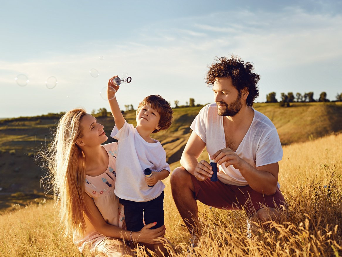 Eine Familie genießt die Freizeit in der Natur, wobei die blonde Mutter und der lockige Vater ihrem kleinen Sohn dabei zusehen, wie er freudig Seifenblasen auf einer sommerlichen Wiese bei Sonnenuntergang bläst.