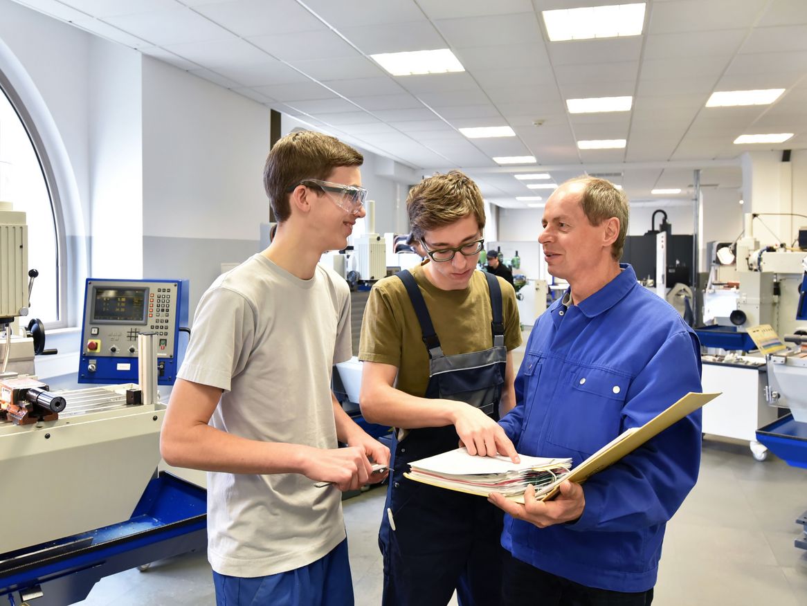Zwei junge Auszubildende in einem technischen Labor sprechen mit einem älteren Lehrer, der ihnen Anleitungen in einem Ordner zeigt, umgeben von Maschinen und Ausrüstung.