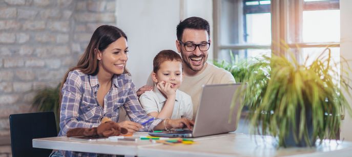 Das Bild zeigt eine Familie mit einer Tochter, die gemeinsam am Esstisch sitzt und auf einen Laptop schaut. Sie befinden sich in einem hellen Raum mit einer Ziegelwand und Fenstern, durch die Tageslicht einfällt.